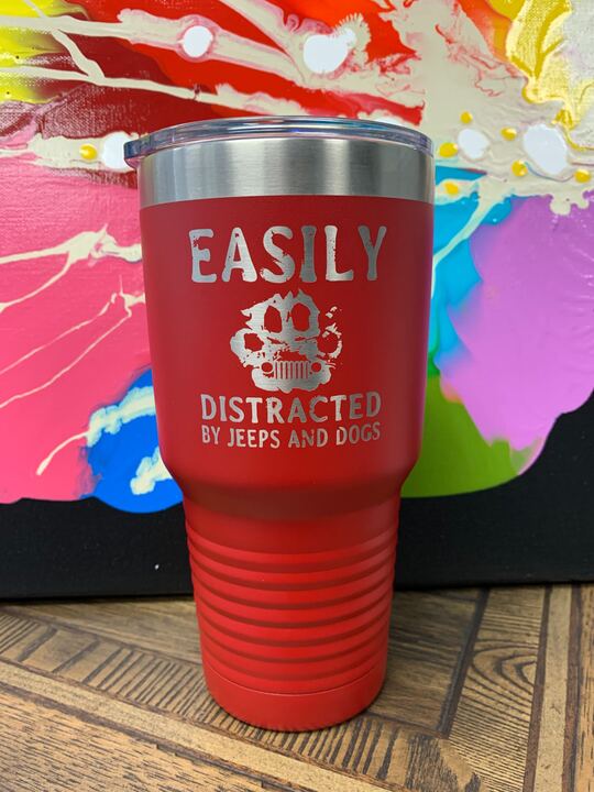Image of an insulated tumbler in Red with metal rim, clear plastic lid, featuring a paw print with a Jeep front outline surrounded by the words "Easily distracted by jeeps and dogs" laser etched in the side of the cup.